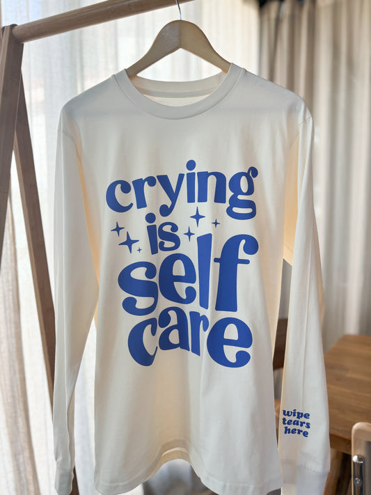 The front of the high-quality long sleeved shirt says "crying is self care" in blue with some sparkles. The wrist of the left sleeve says, also in blue, "wipe tears here".
