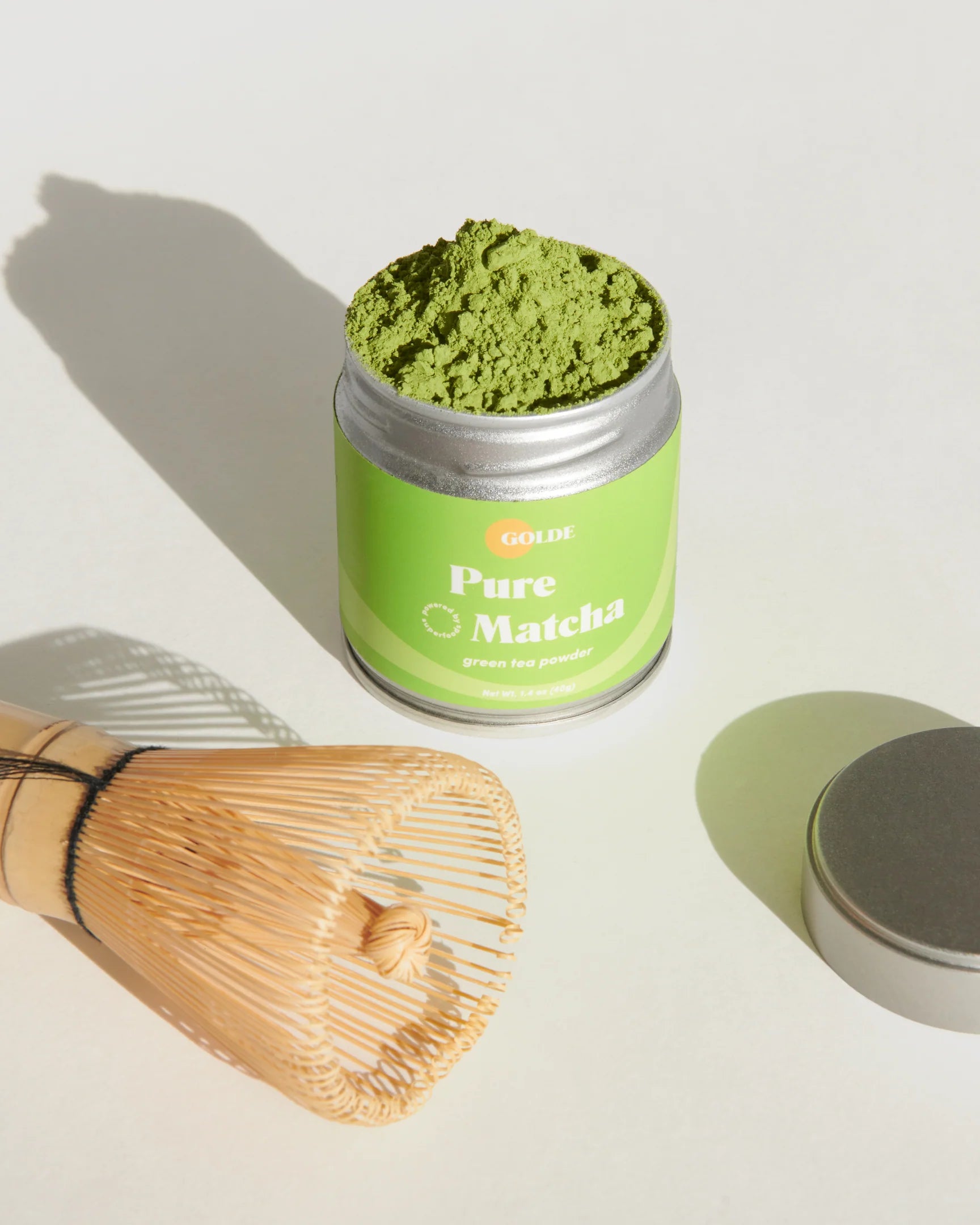 A high-quality photo of the delectable matcha overflowing from the tin, alongside the cap for the tin and the sustainable bamboo whisk.