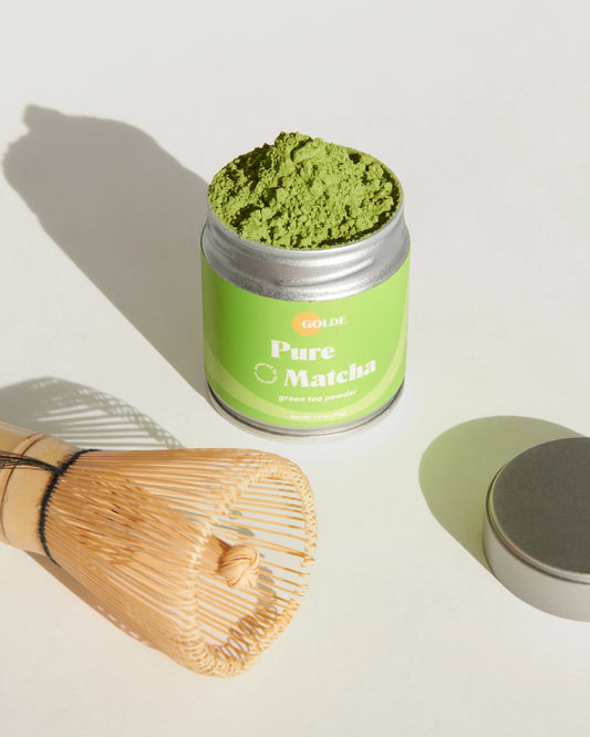 A high-quality photo of the delectable matcha overflowing from the tin, alongside the cap for the tin and the sustainable bamboo whisk.